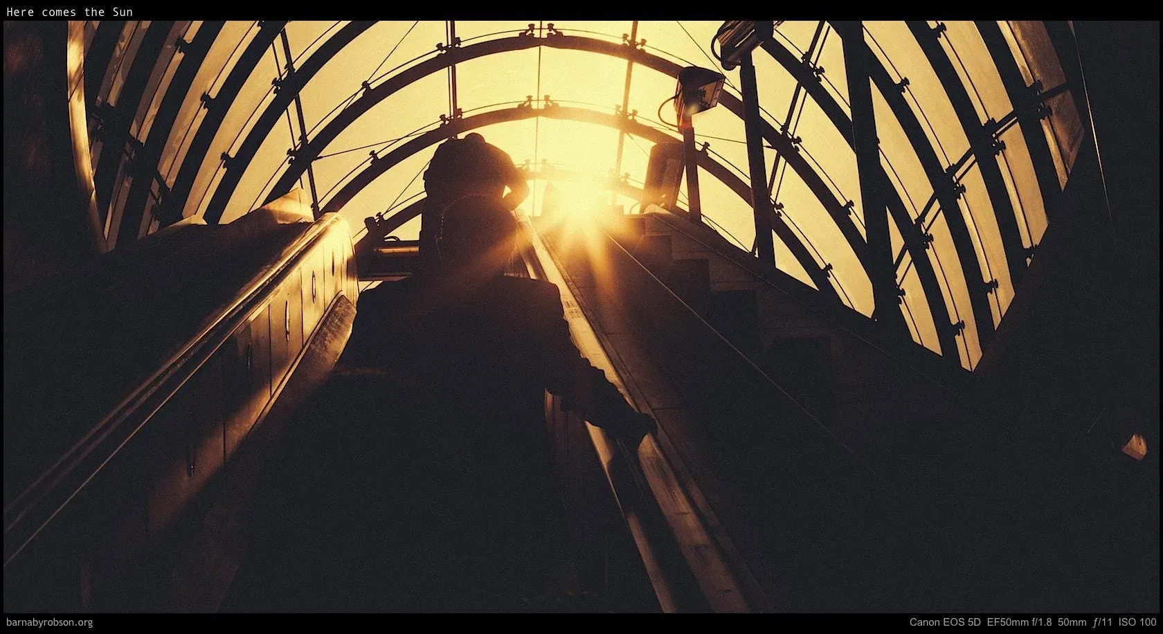Commuters descending into a London station as low sun bursts through the ironwork, black and white.