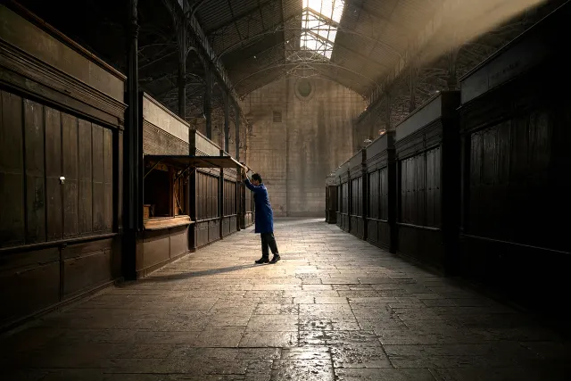 A trader raising the first shutter in a vast empty Victorian market hall at dawn, light falling from the roof glass.