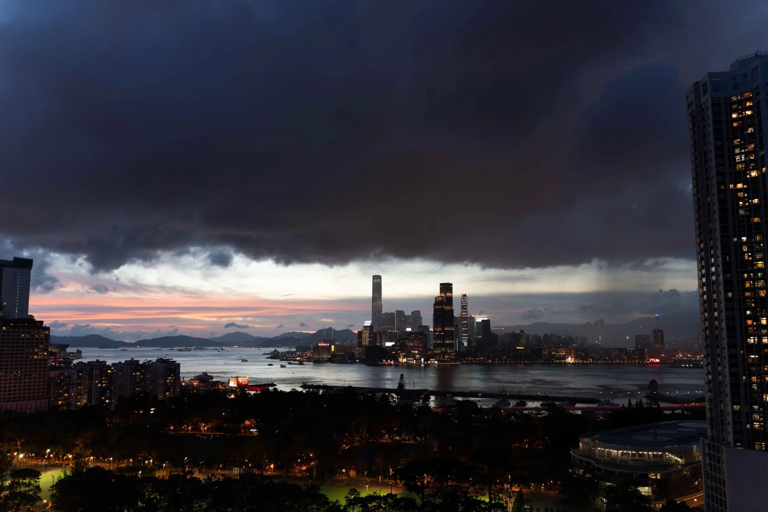 Illuminated city skyline at dusk with harbour, dark clouds, and a colourful sunset.