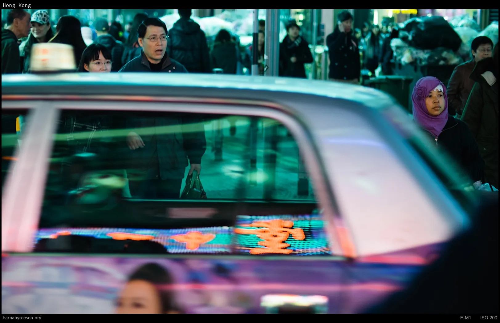 People on a busy street, a taxi in foreground reflects neon signs.