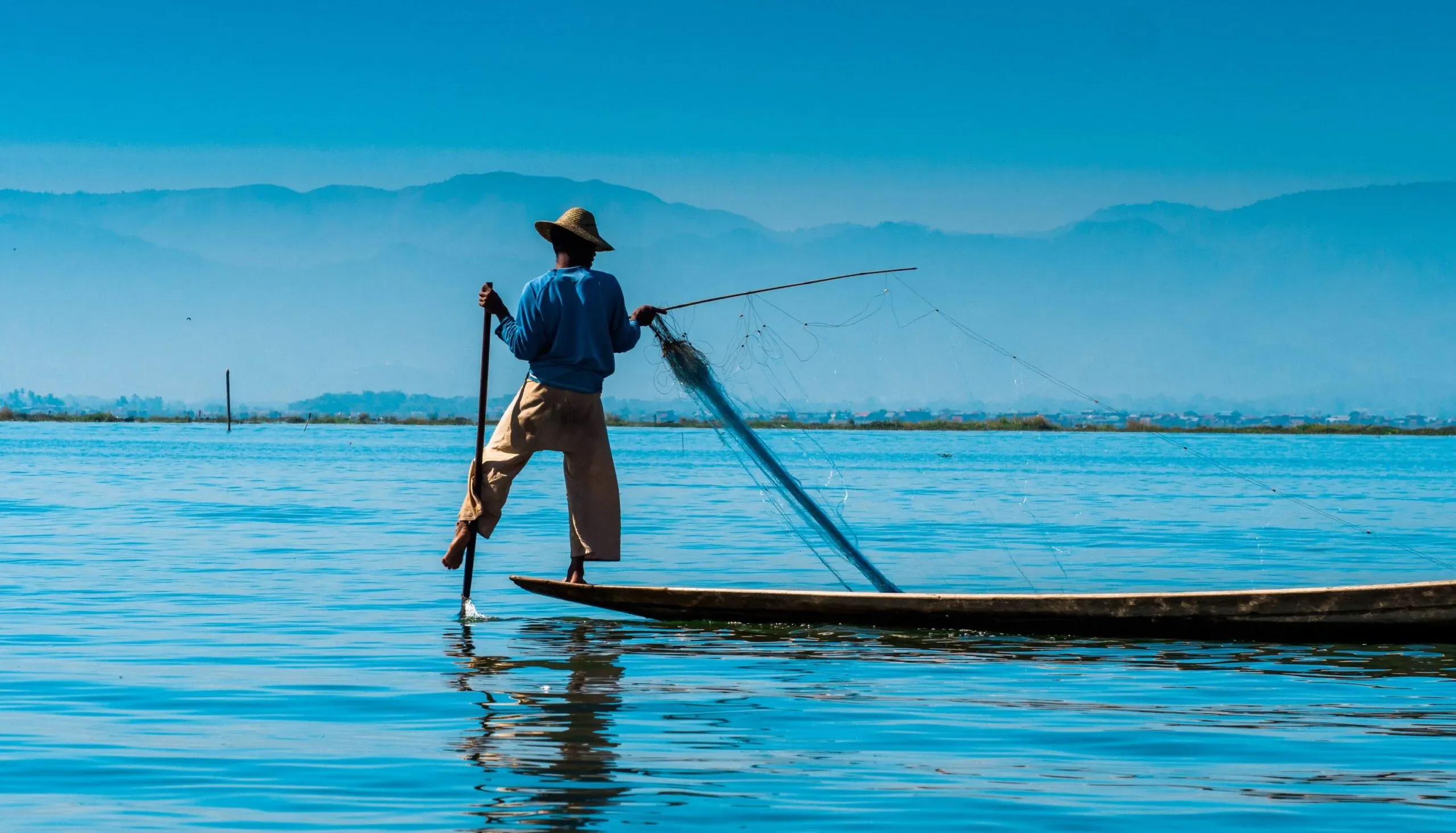 An Intha fisherman rowing one-legged on Inle Lake, net in hand against blue haze