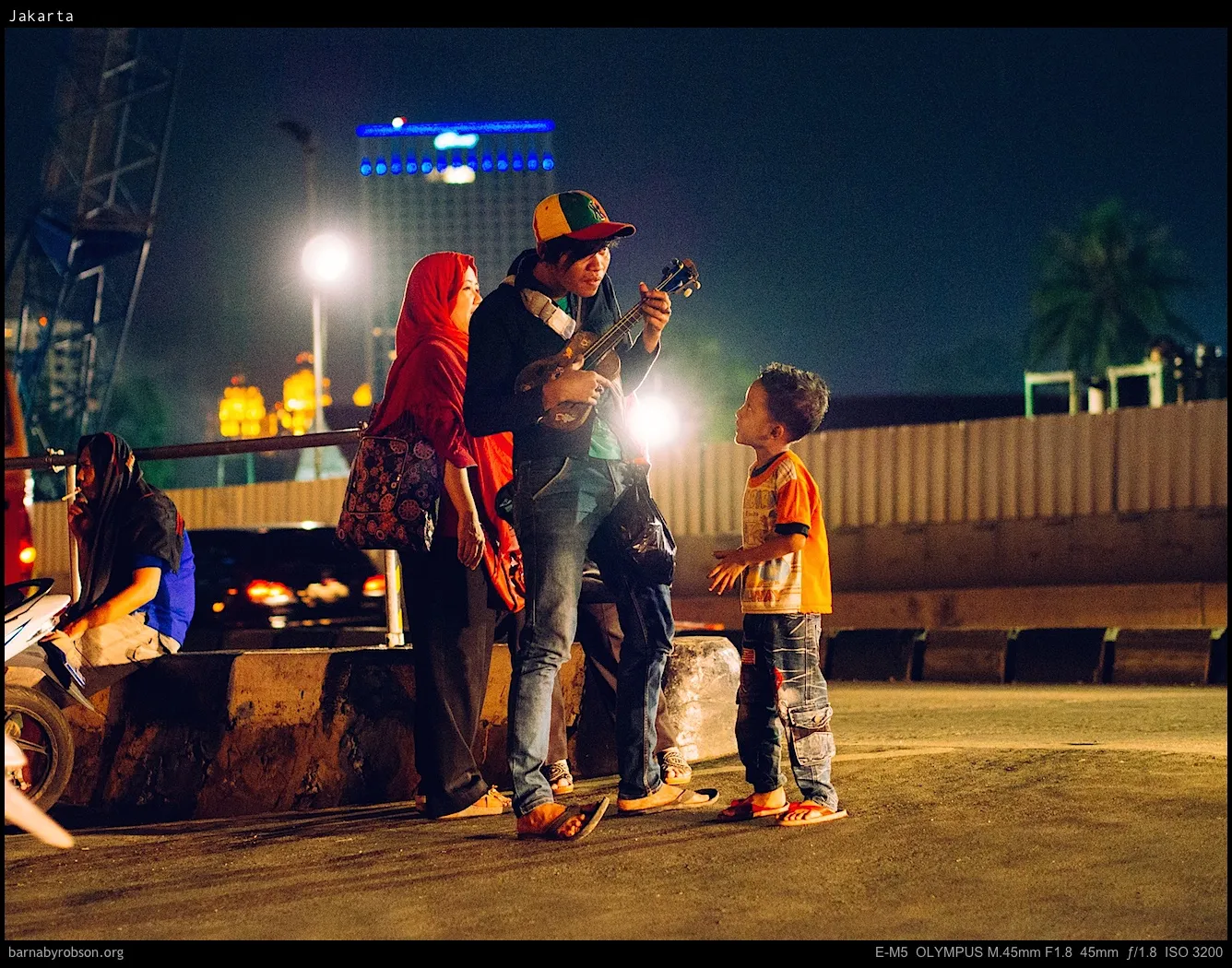 Man in cap plays ukulele for boy and woman on night street.