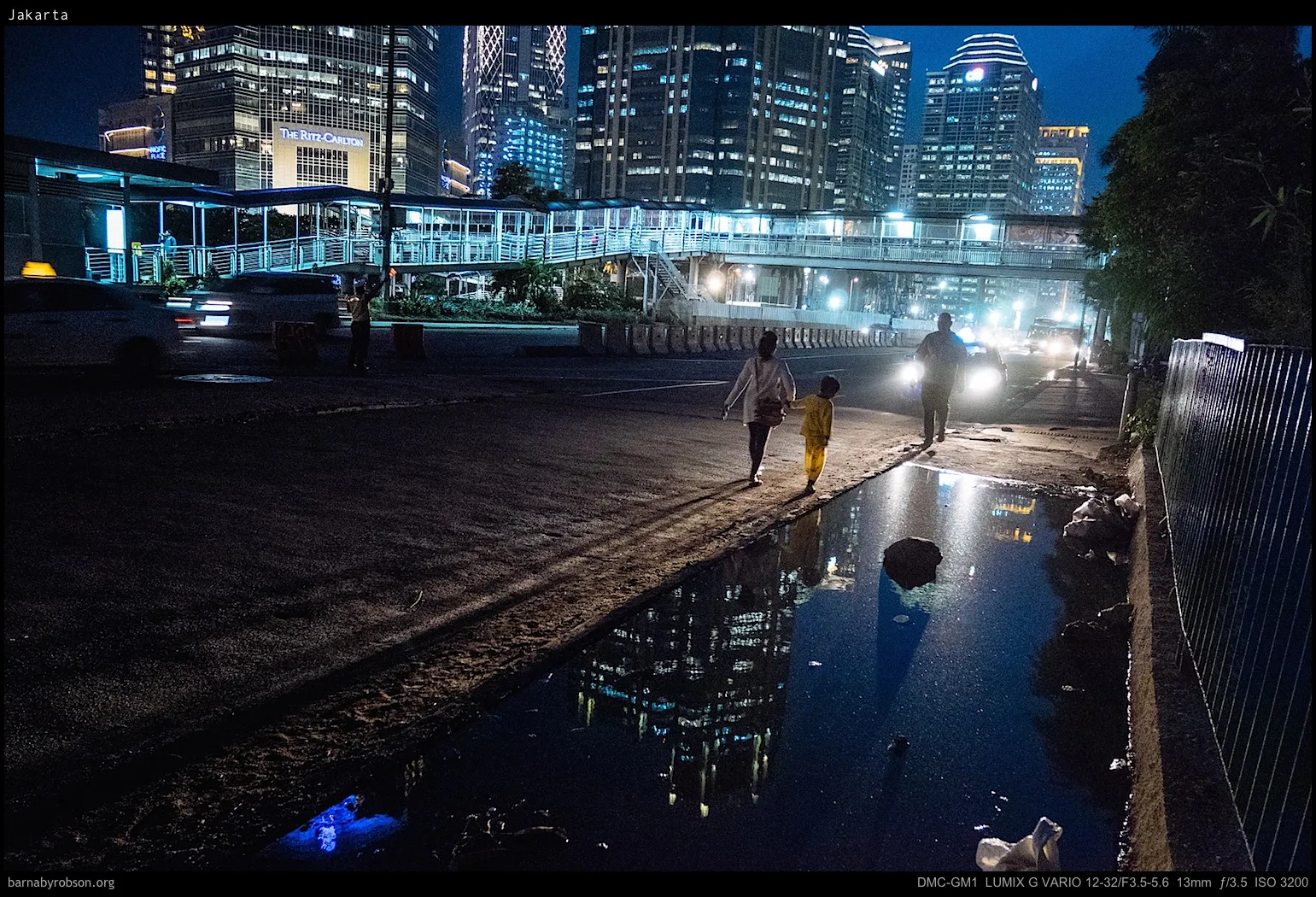 Night city street with pedestrians, cars, and illuminated buildings reflecting in a puddle.