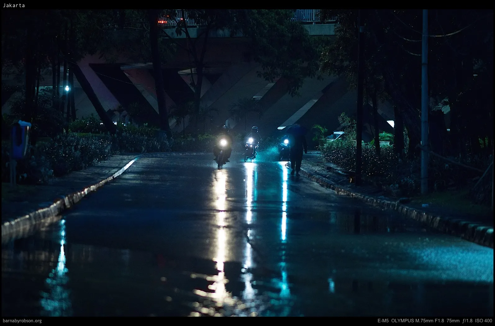Dark, wet street with motorbikes and pedestrian under umbrella.