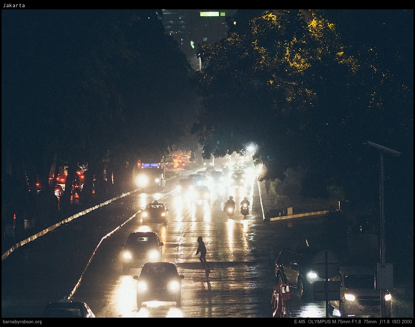 Wet night road with cars, motorbikes, and a person crossing.