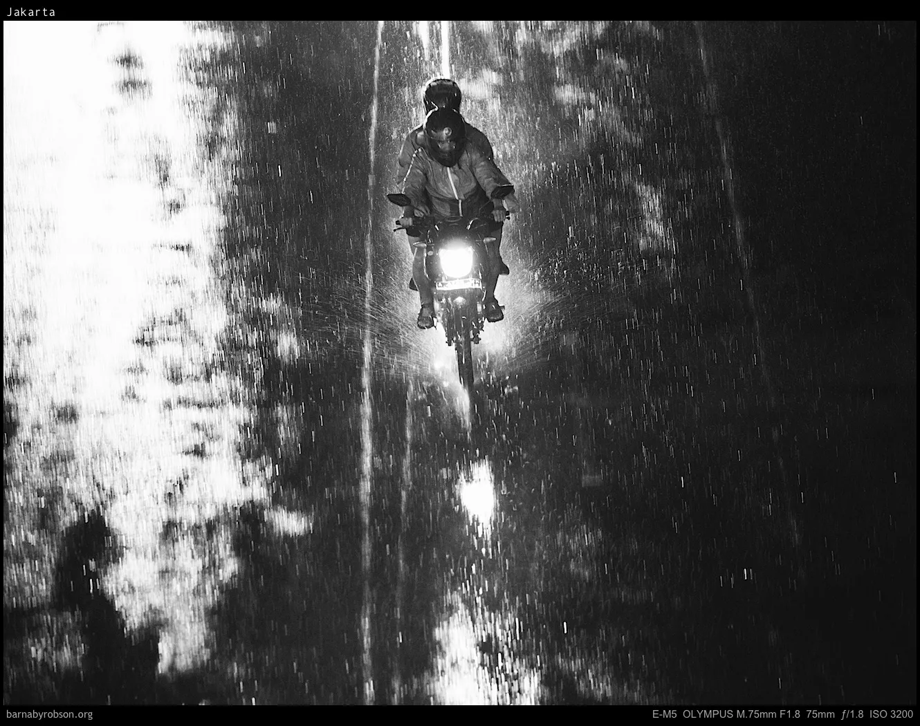 Two people on a motorbike ride through pouring rain at night.