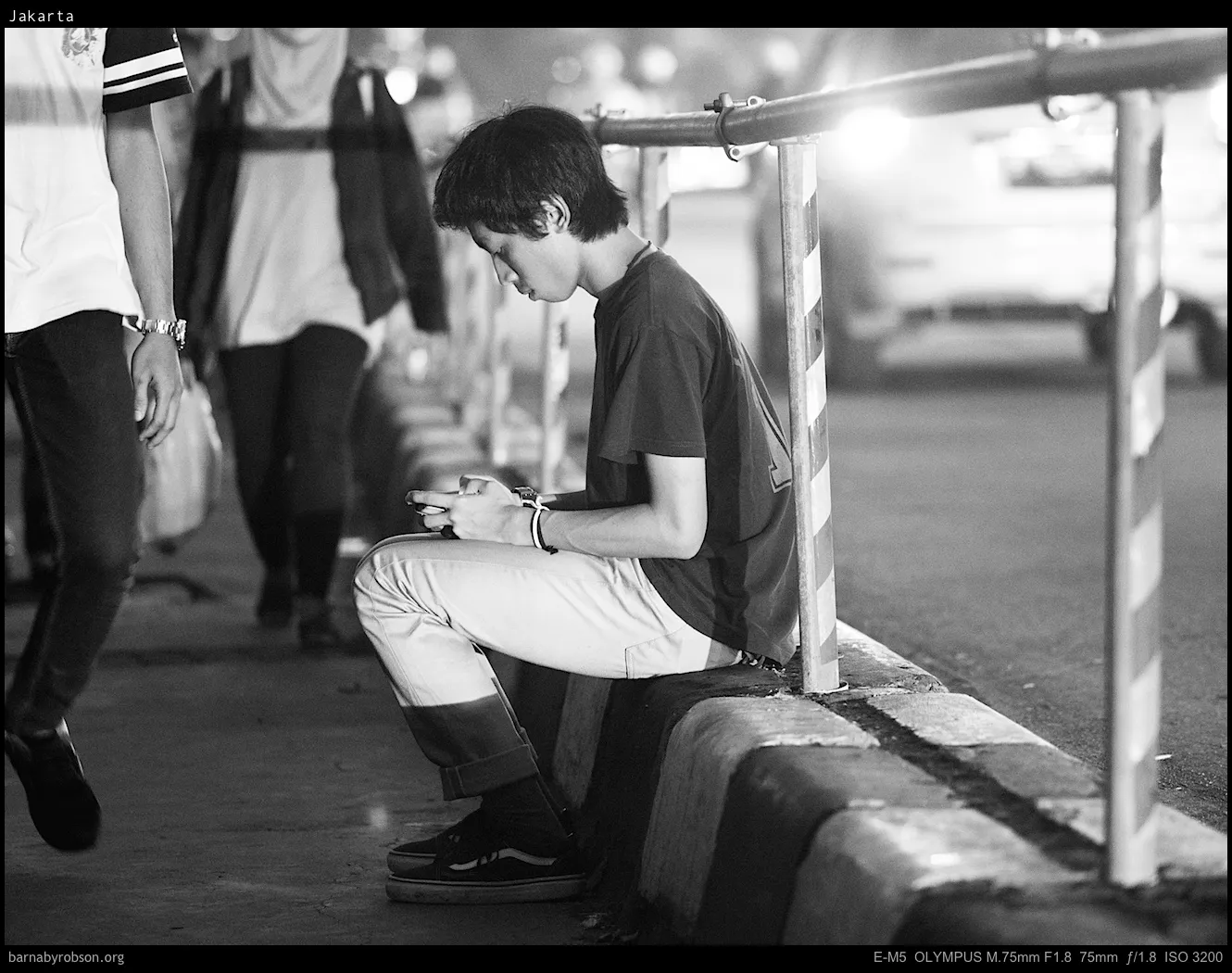 Young person sits on kerb, looking at phone, beside a striped barrier.