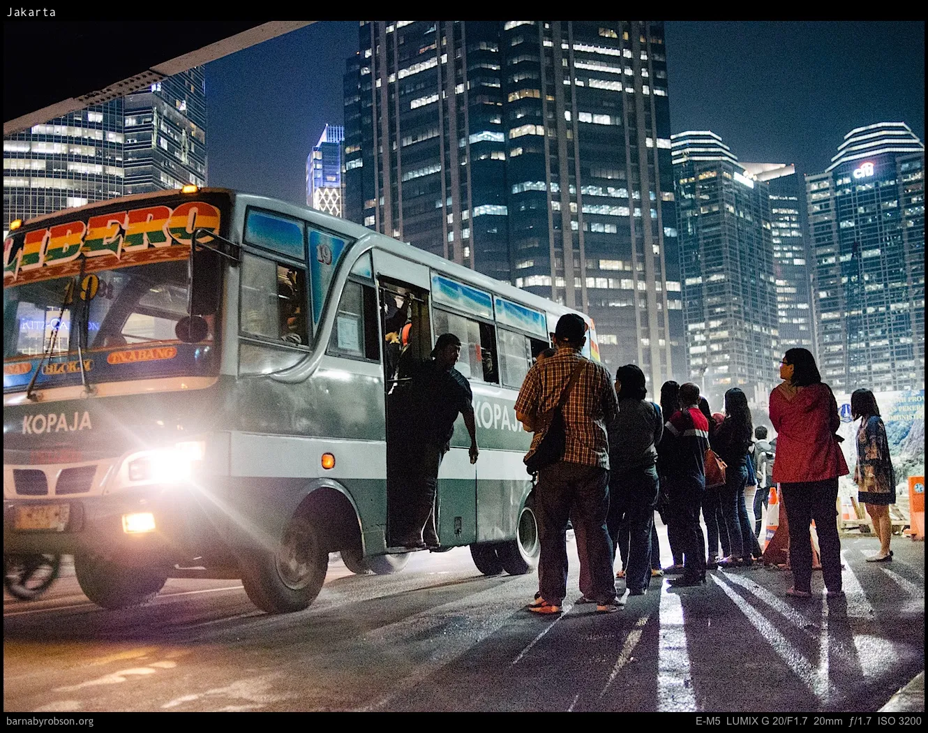 Bus with bright headlights, people on street, illuminated city buildings at night.