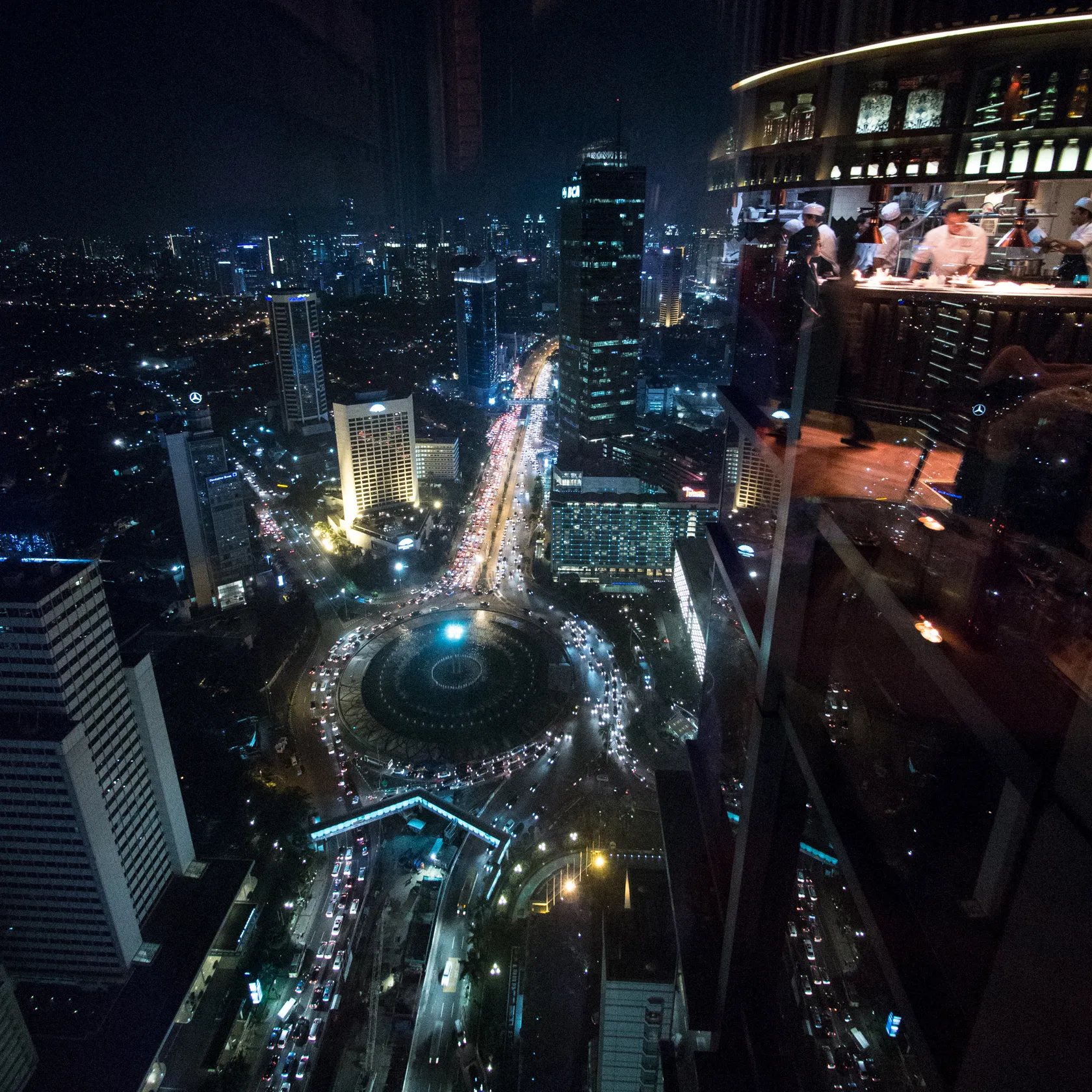 Night city view with busy traffic, a fountain roundabout, and bar reflection.