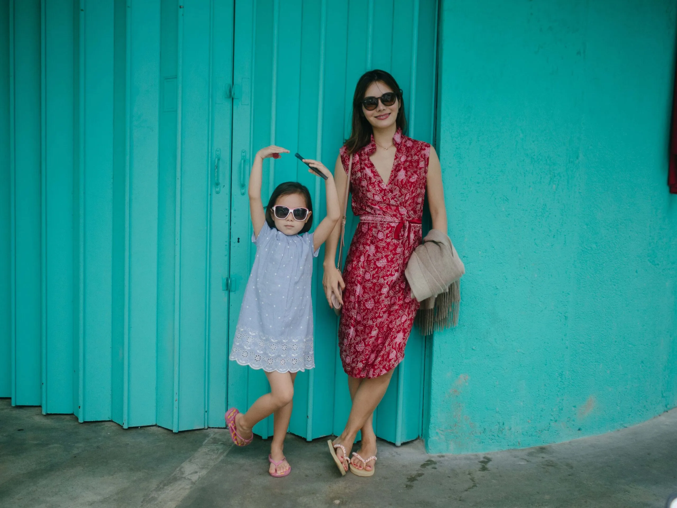 Woman and girl in sunglasses against a teal wall, girl with arms raised.