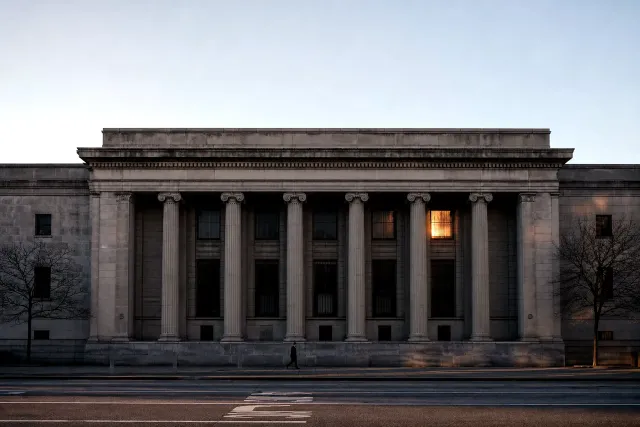 The stone facade of a grand central bank at cold dawn, one commuter passing small beneath the columns, a thin band of sunrise caught in a high window.