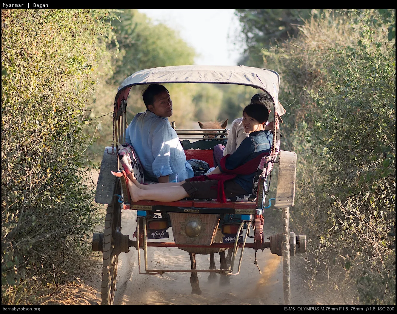 Two people riding in a horse-drawn cart on a dusty track.