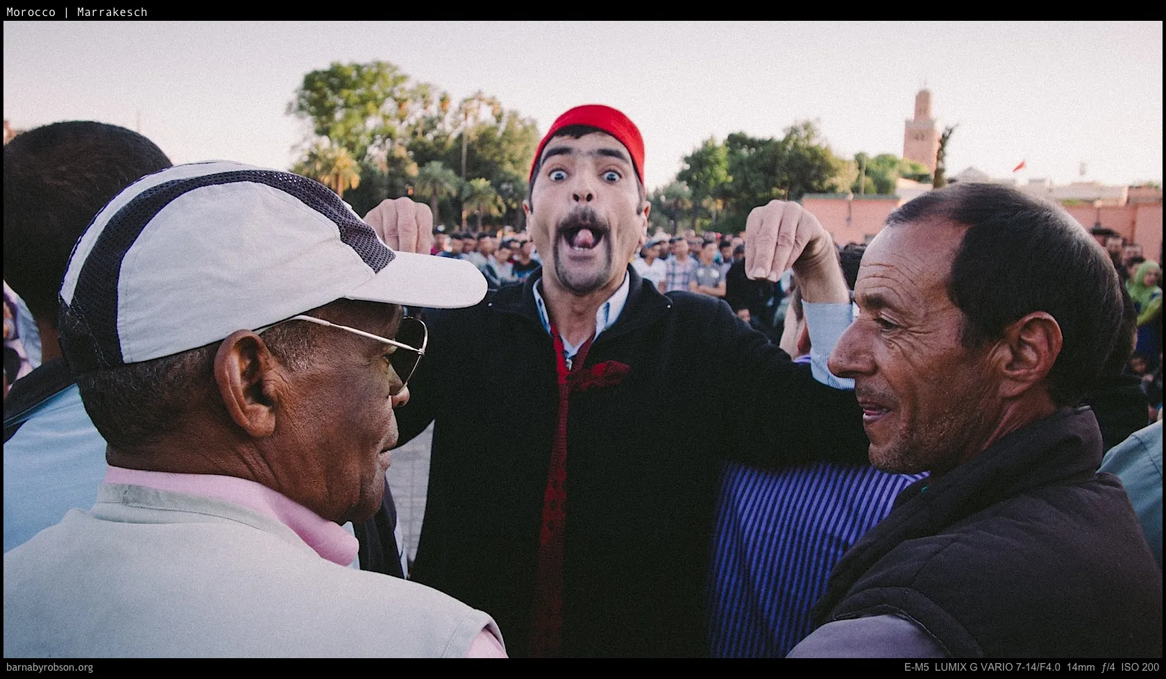 Man in red hat pulls funny face for two men amidst a crowd.