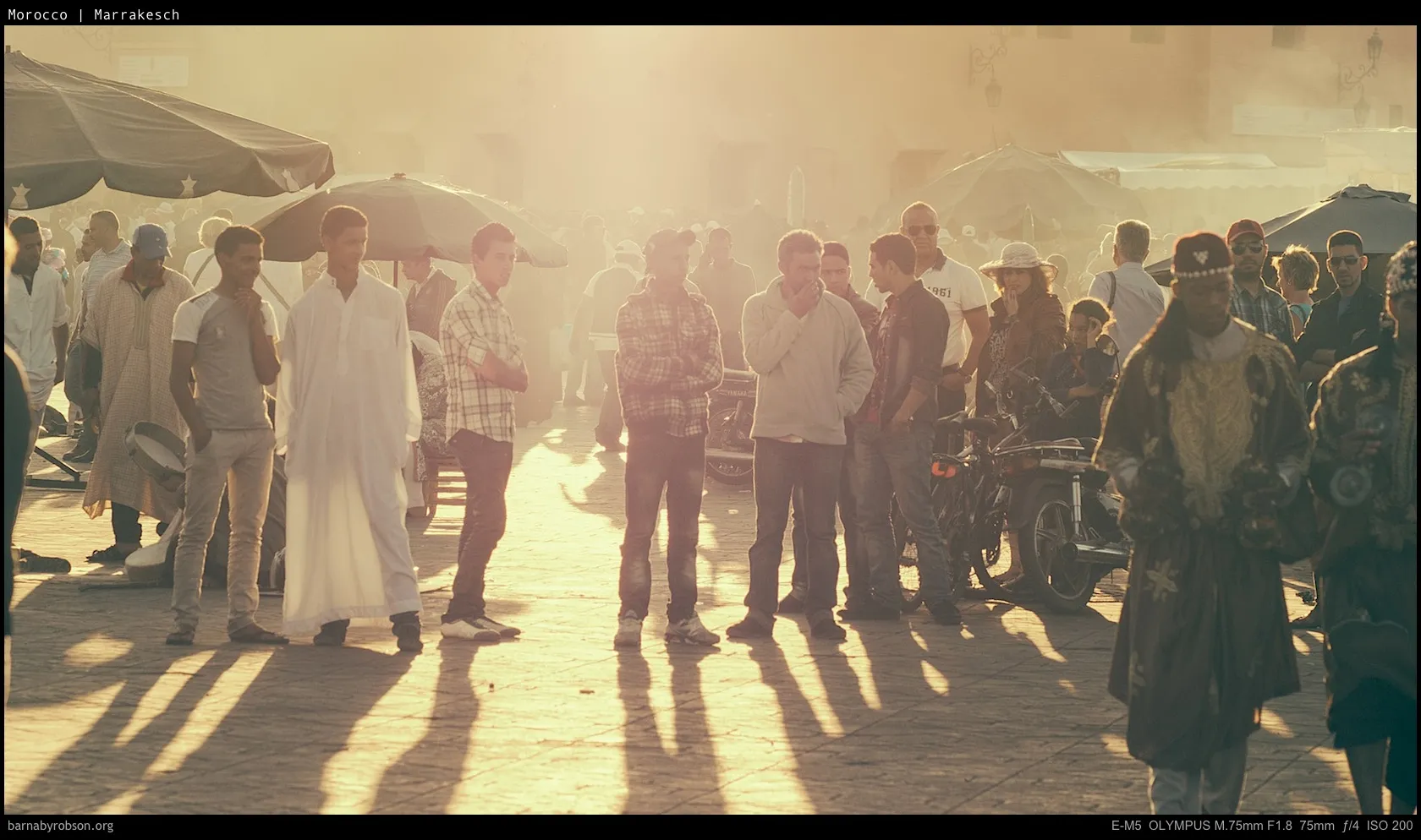 Busy Marrakesh market scene with people, traditional dress, and long shadows.
