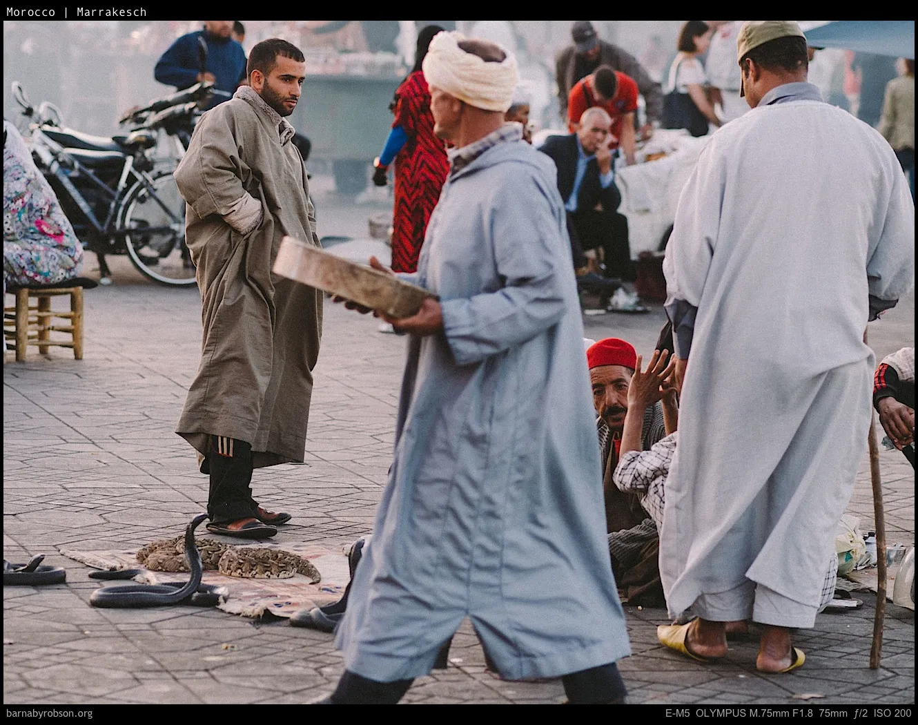 Men in traditional robes, a snake charmer with snakes on a paved street.
