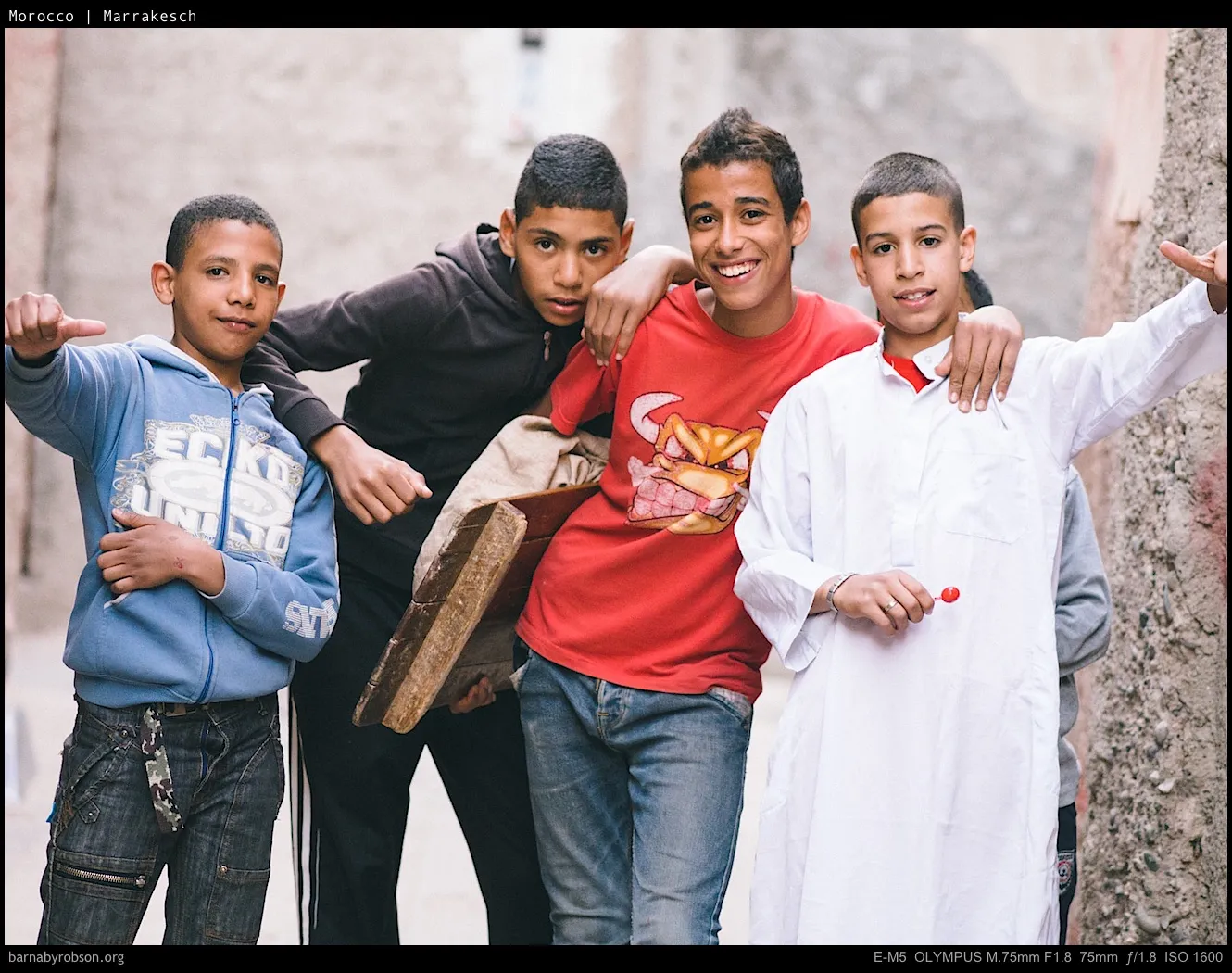 Five boys posing in a narrow street, one holding a red lollipop.