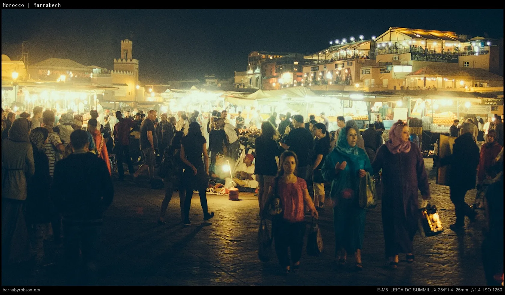 Crowded night market with glowing stalls, people walking, and a distant tower.