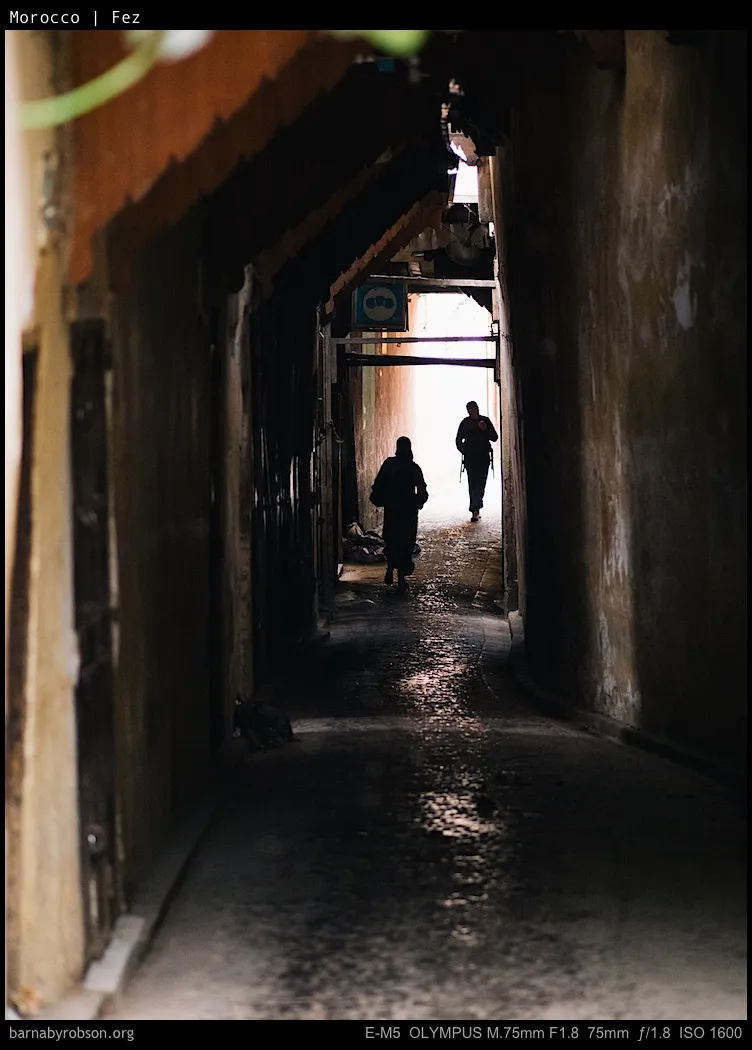 Silhouetted figures walk down a narrow, wet alley towards a bright exit.