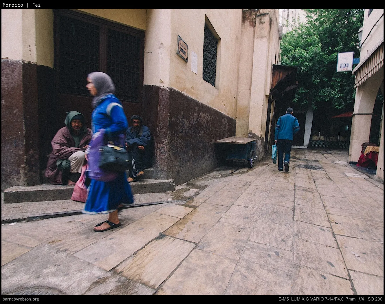 Woman walks past men sitting on steps in a narrow, old street.