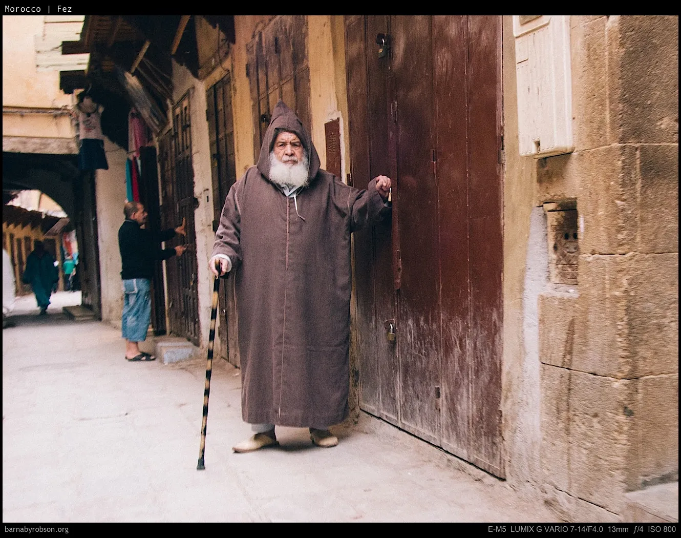 Elderly man in brown robe with white beard and walking stick in a narrow street.