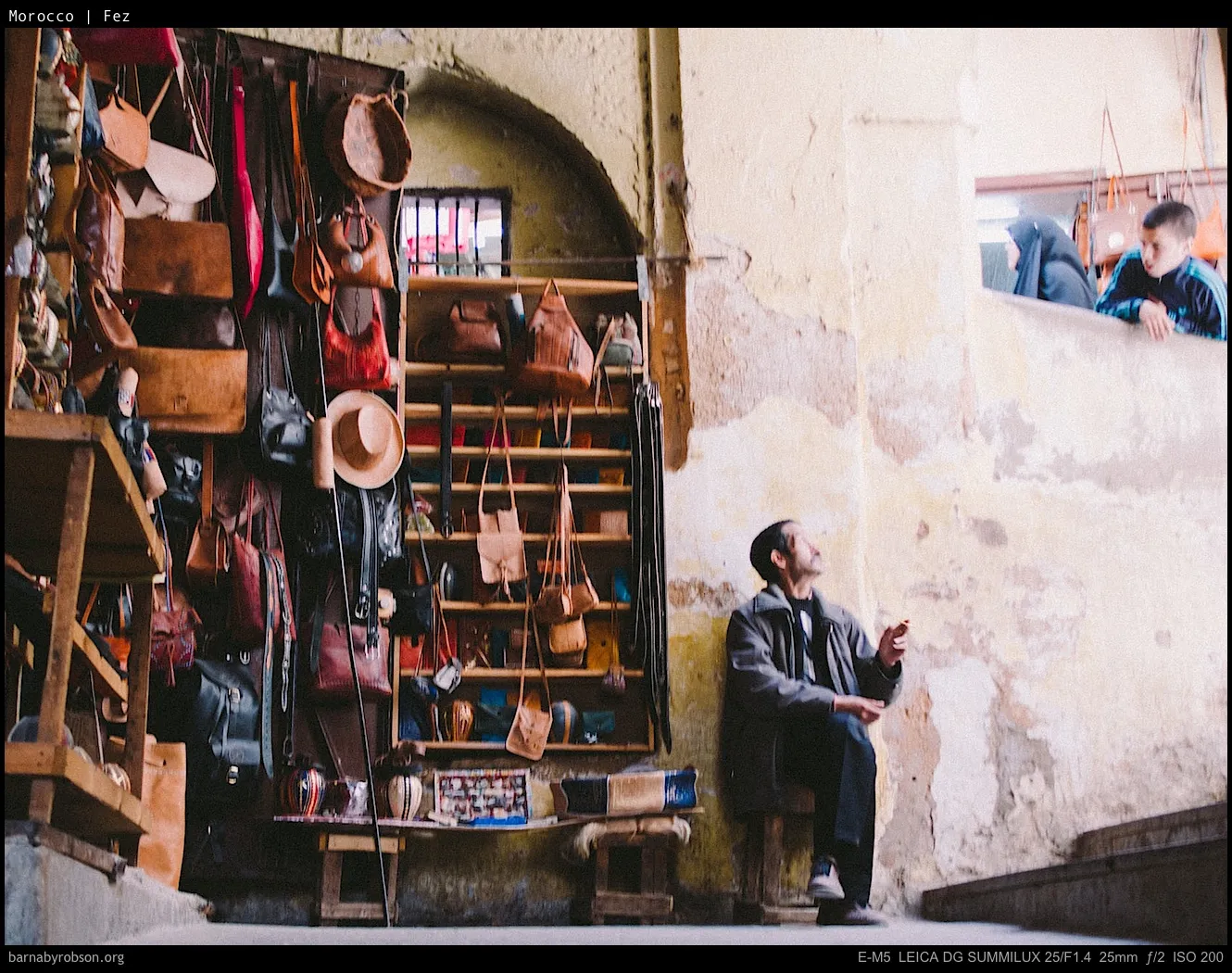 Man sits in leather goods shop; two people peer from upper opening.