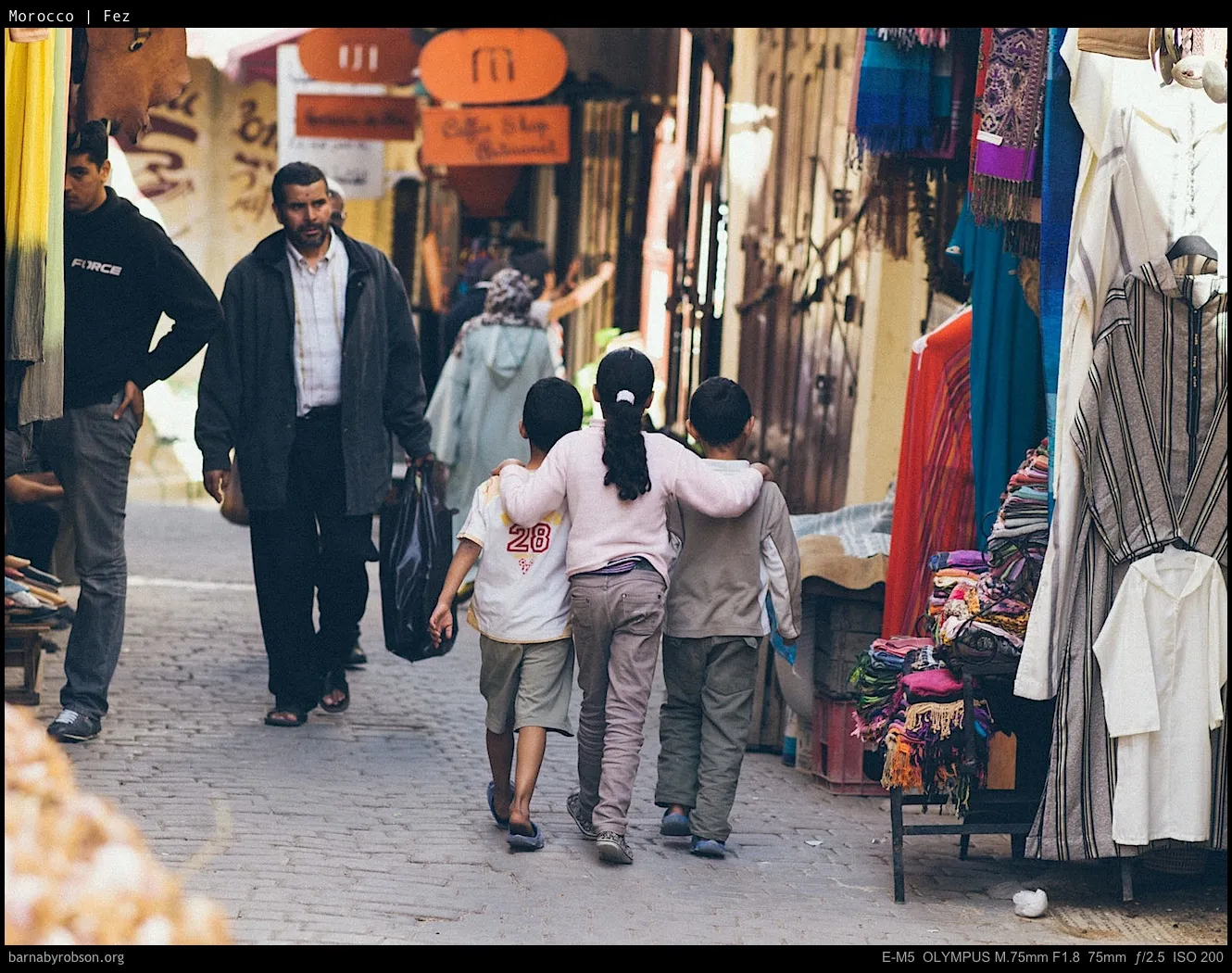 Children walk arm-in-arm down a narrow market street with people and textile stalls.