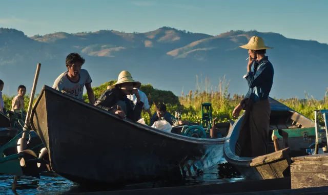 Women in conical hats crossing Inle Lake by wooden boat, mountains behind.