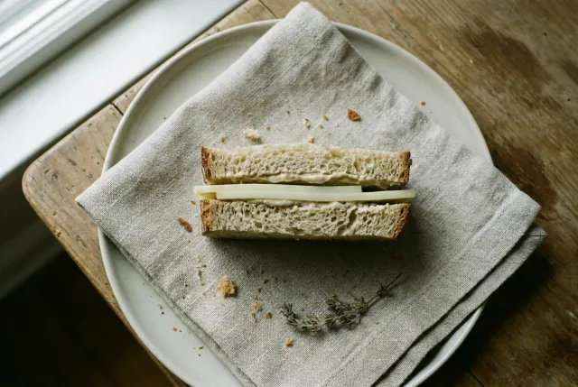 A simple handmade sandwich cut in half on a linen napkin and pale plate in soft window light, its layers on show — a nod to the human-AI sandwich: you frame the task, the machine fills the middle, you finish it.