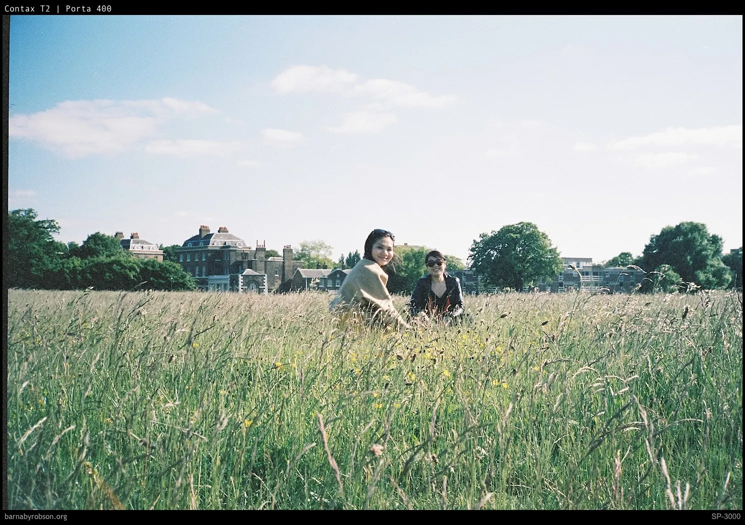 Two smiling women sit in a tall grassy field with buildings and trees behind.