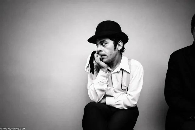 Ian Dury sitting against a pale studio wall in a bowler hat and braces, chin resting on his hand, gloves hanging from his fingers.