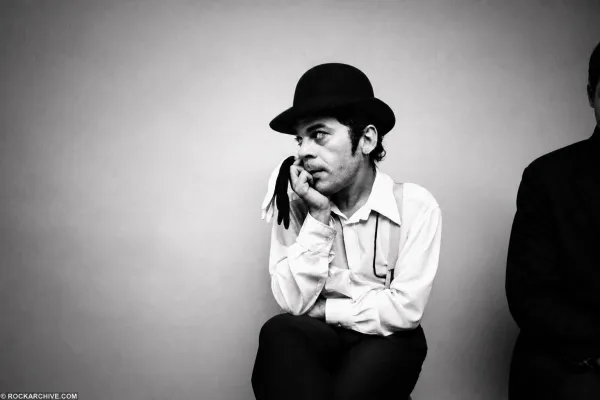 Ian Dury sitting against a pale studio wall in a bowler hat and braces, chin resting on his hand, gloves hanging from his fingers.