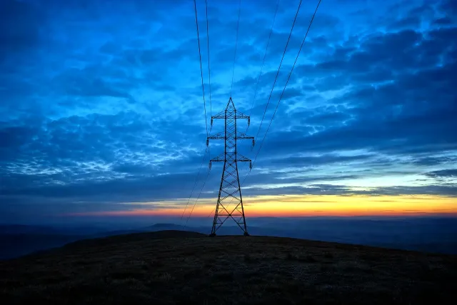 A steel transmission pylon on an open ridge at blue hour, power lines receding into haze beneath a deep blue sky with one warm band of dusk light on the horizon.