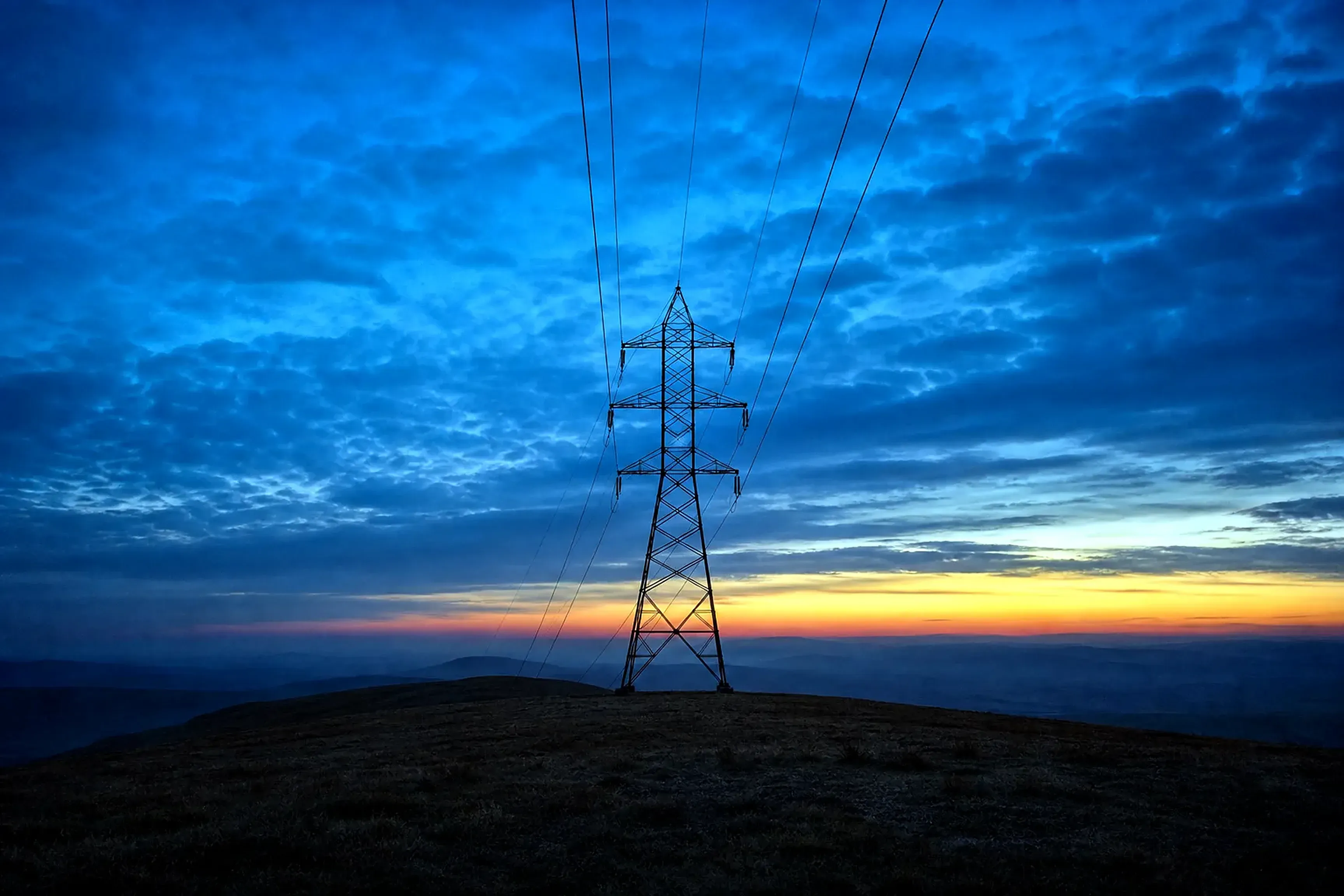 A steel transmission pylon on an open ridge at blue hour, power lines receding into haze beneath a deep blue sky with one warm band of dusk light on the horizon.