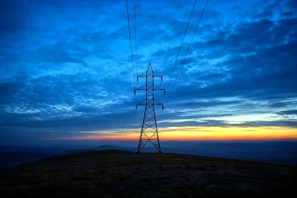 A steel transmission pylon on an open ridge at blue hour, power lines receding into haze beneath a deep blue sky with one warm band of dusk light on the horizon.