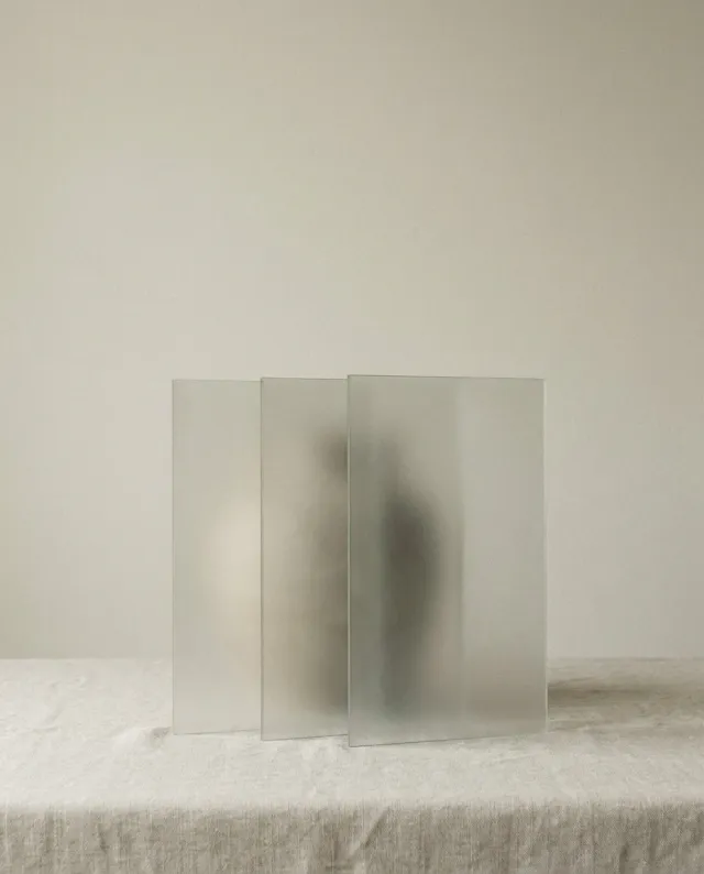 A minimal still-life photograph of three tall frosted translucent panels standing and gently overlapping on a pale surface in soft light — knowledge, power and money.