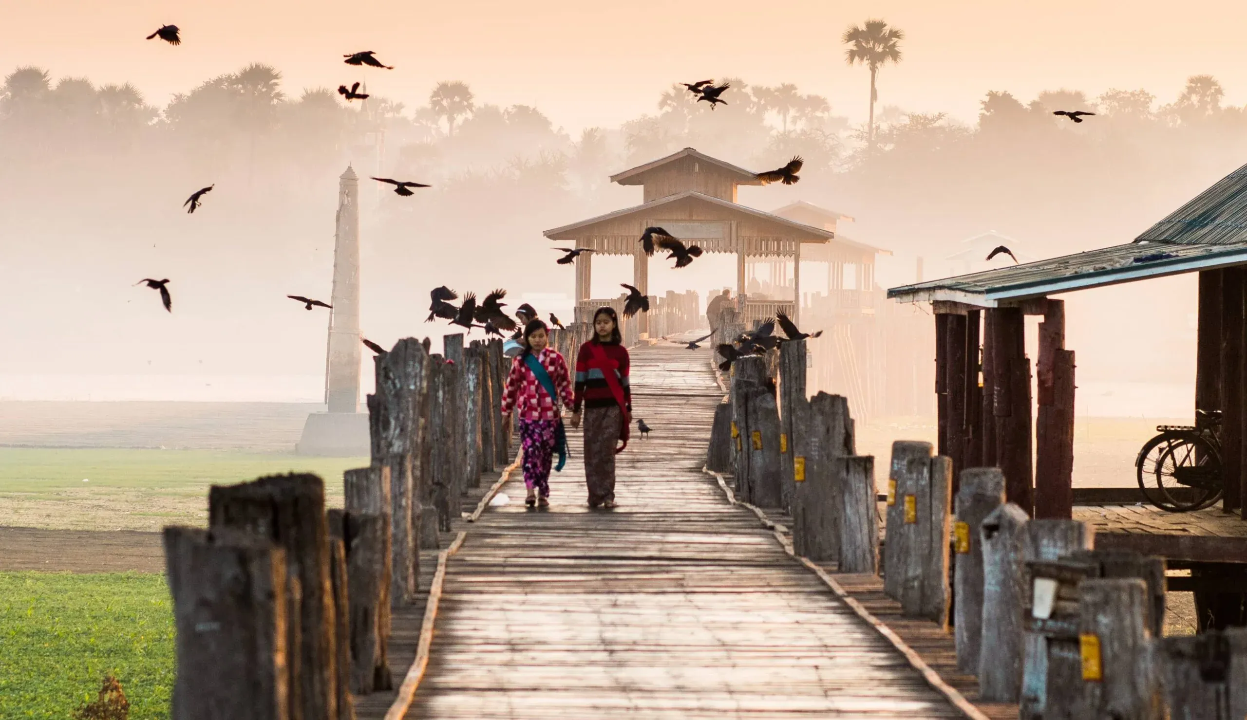U Bein Bridge at dawn: two girls on the teak walkway, crows wheeling through the mist