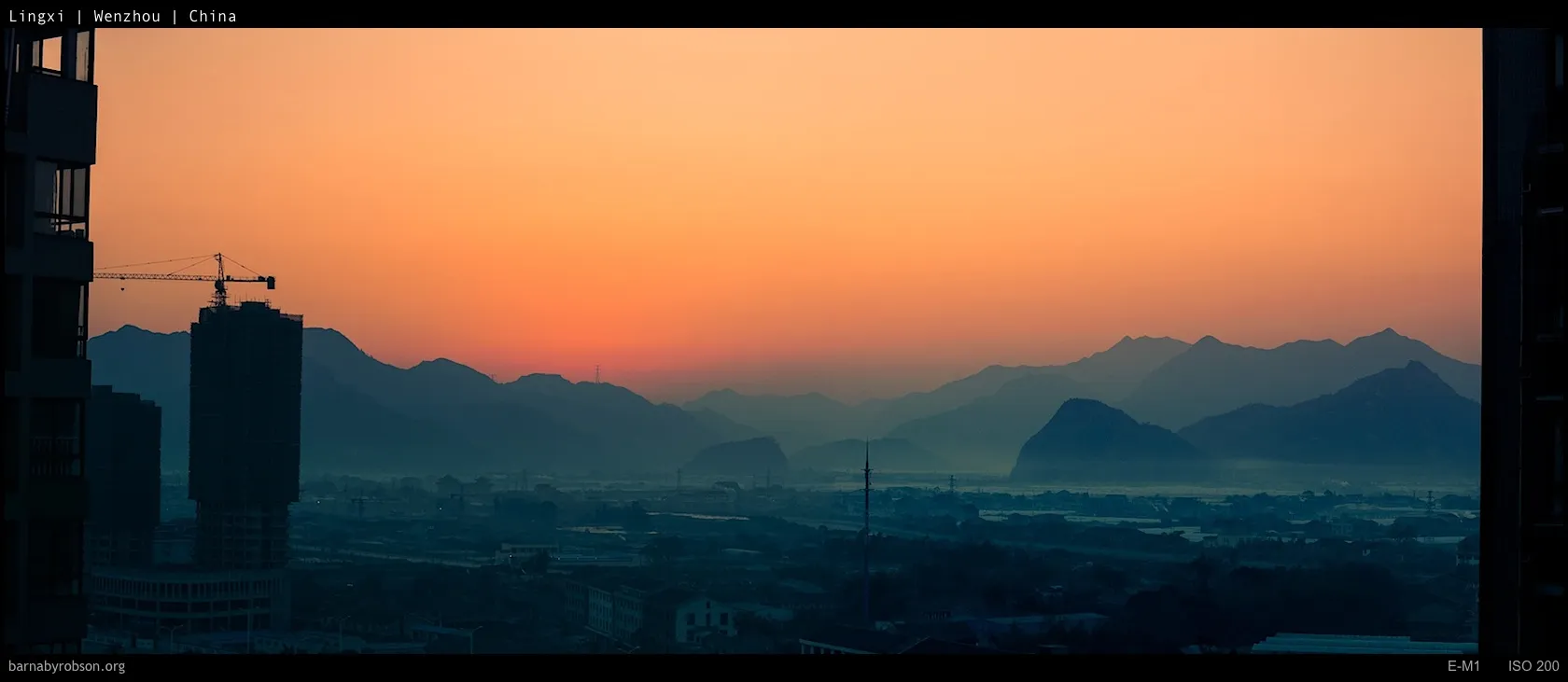 Orange sunset sky above hazy mountains, city, and a construction crane.