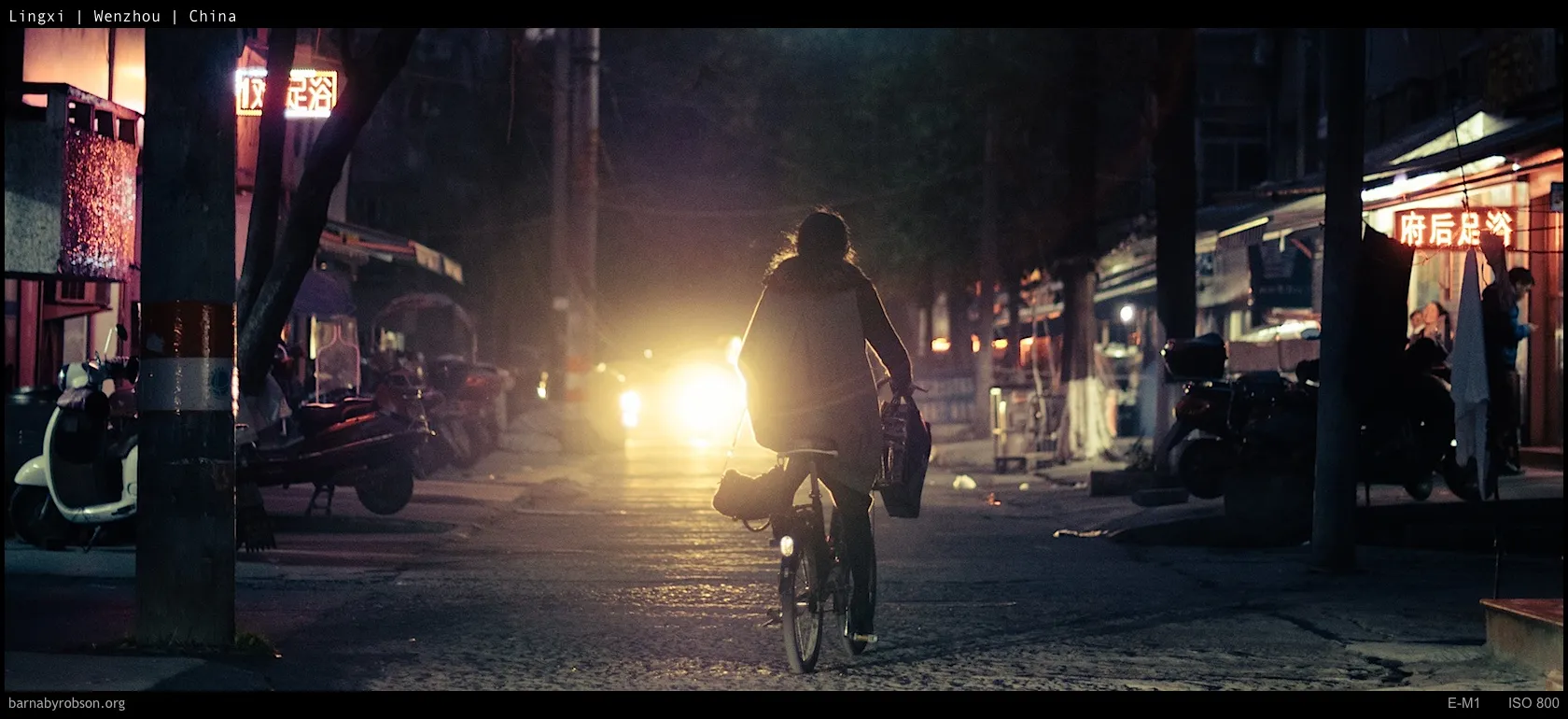 Night street scene with a person cycling towards bright car headlights.