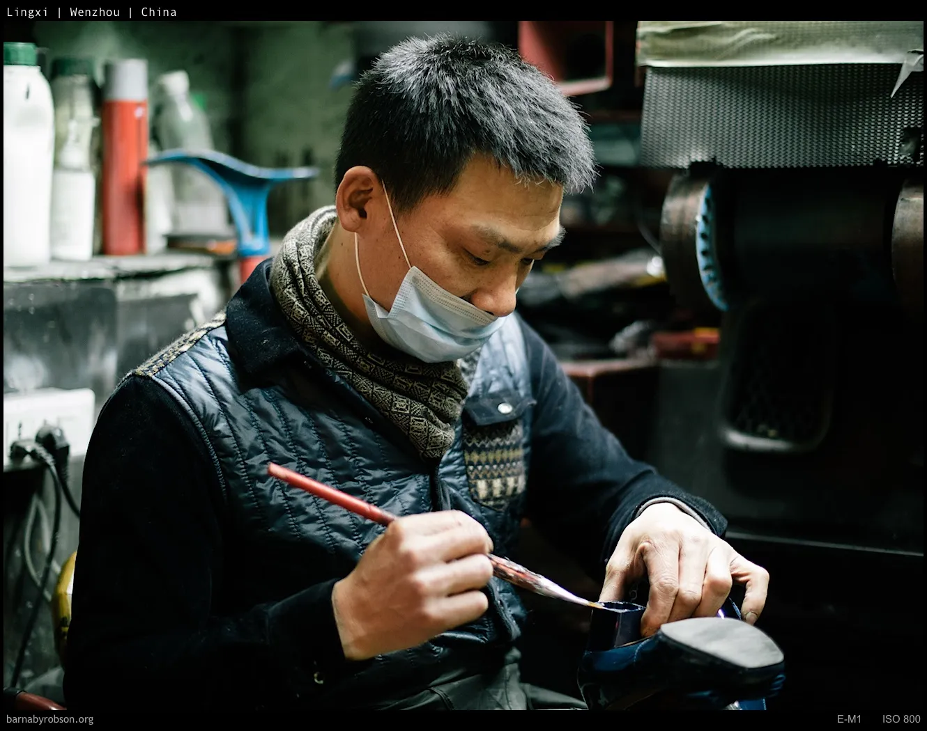 Masked man carefully brushes a dark blue shoe in a workshop.