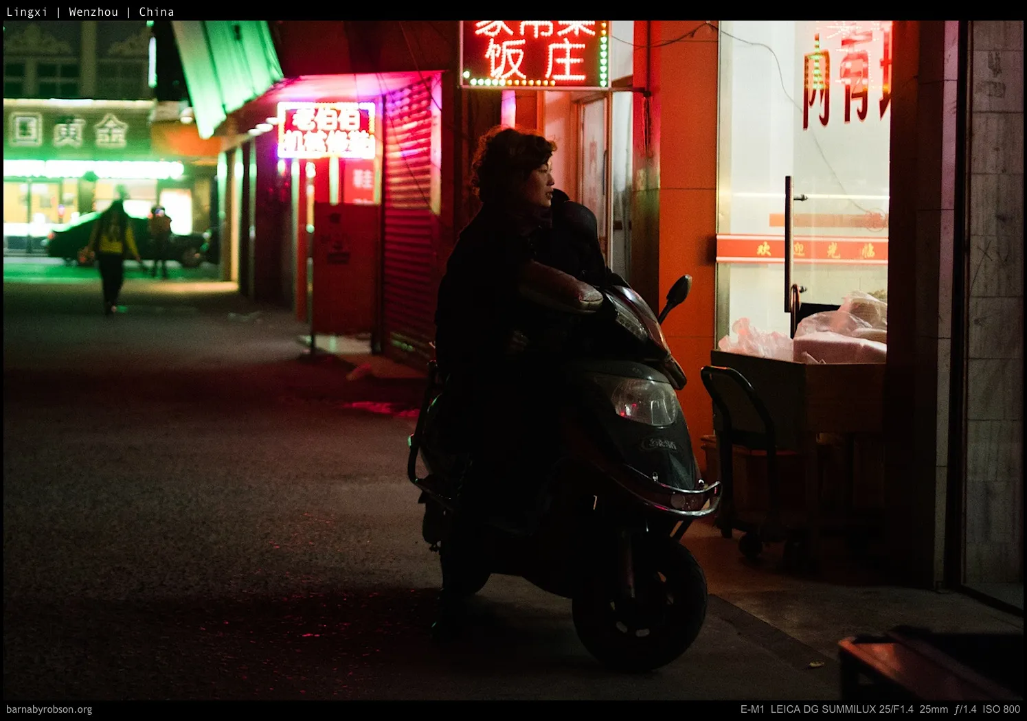 A person on a scooter silhouetted against neon signs on a Chinese street at night.