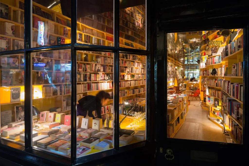 The warmly lit window of a densely stocked independent bookshop at night