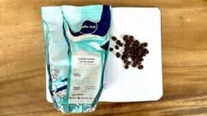 Roasted coffee beans next to a dotted notebook on a wooden surface, with a bag of Arabica Sundan Giling Basah coffee from Kafe Inc, highlighting premium coffee selection.