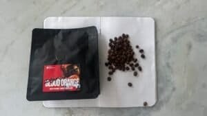 Rich coffee beans and a bag of Space Roastery’s Blood Orange chocolate nibs on a white textured surface.