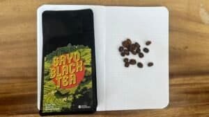 Rich black coffee beans on an open notebook with dotted pages, alongside a colourful "Gayo Black Tea" packet on a wooden surface, representing artisanal coffee and tea products.