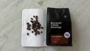 Rich, dark coffee beans spilled on a notebook next to a bag of ブラックリストコーヒーロースターズ Papayo Bourbon coffee, highlighting speciality beans for espresso lovers.