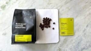 Bright yellow and black coffee packaging with roasted coffee beans on a white dotted notebook, showcasing premium Indonesian Blacklist coffee beans, highlighting speciality coffee beans and sustainable sourcing.
