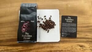 Rich roasted coffee beans from Barnaby Robson’s The Forbidden Flower blend on a dotted notebook, beside the product packaging and an informational card, highlighting artisanal coffee, single origin, and flavour profiles.