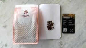 Freshly roasted Malabar Peeberry coffee beans on a dotted notebook with a Smoking Barrels craft coffee bag and a QR code coffee card, perfect for coffee enthusiasts and artisanal brewing.