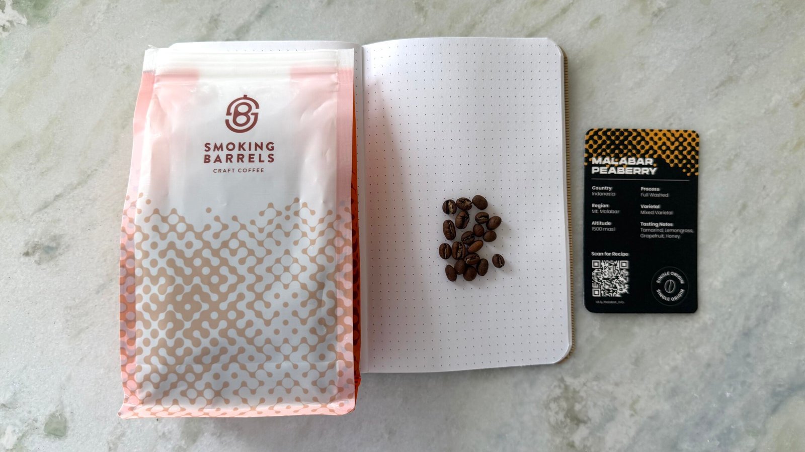 Freshly roasted Malabar Peeberry coffee beans on a dotted notebook with a Smoking Barrels craft coffee bag and a QR code coffee card, perfect for coffee enthusiasts and artisanal brewing.