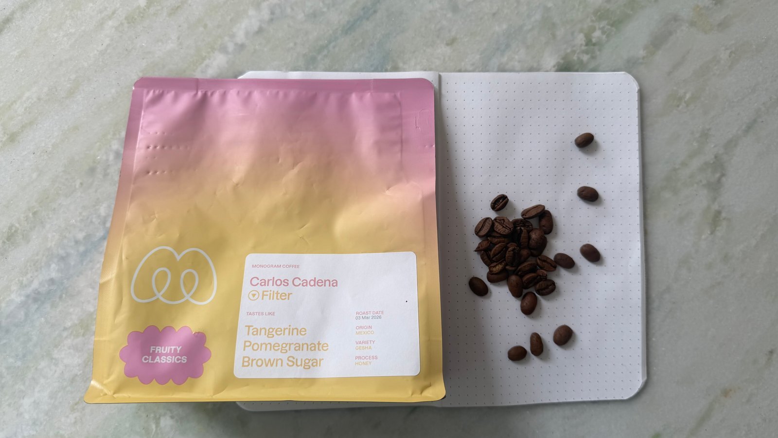 Close-up of a bag of Carlos Cadena Filter Tangerine Pomegranate Brown Sugar with some coffee beans on a white notebook.